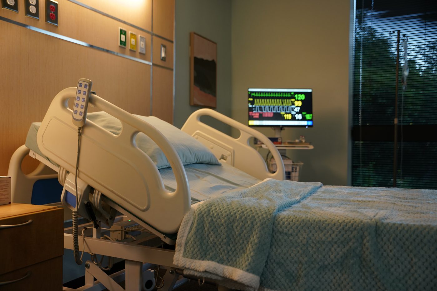 Hospital standing set: adjustable bed, vitals monitor, wood-panel headwall and a window with blinds looking out to trees