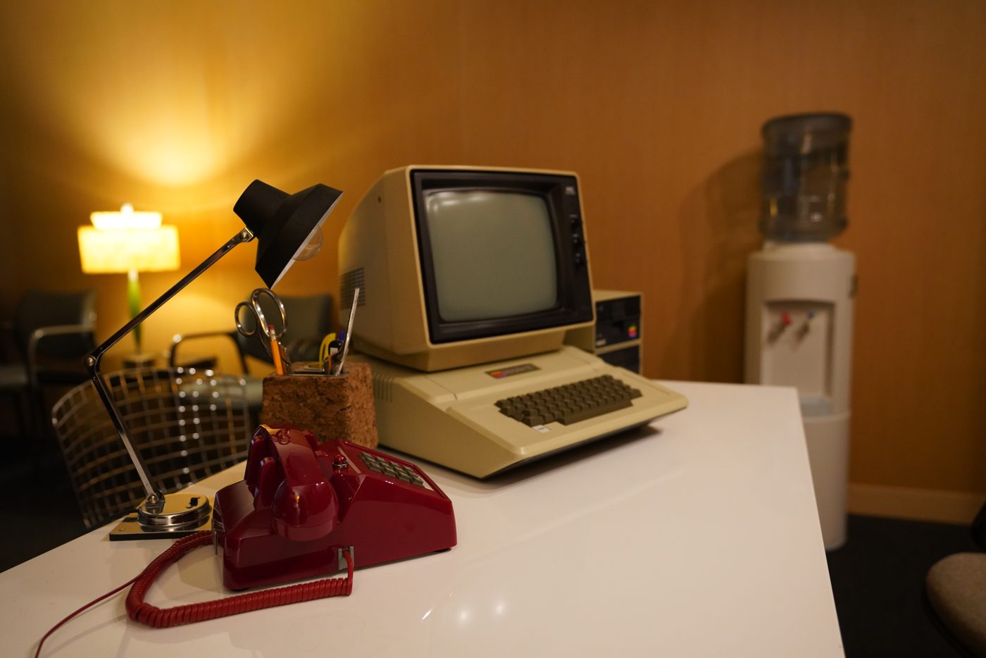 Retro office standing set: vintage computer, red desk phone and desk lamp on a white desk, with a water cooler and warm wood walls behind